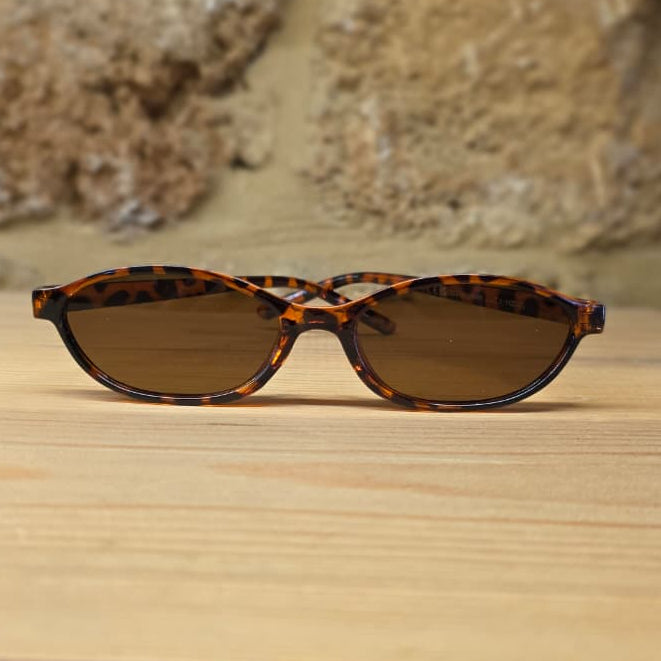 Oval sunglasses with a brown and black tortoiseshell patterned frame and dark brown lenses, resting on a light wooden surface with a blurred stone wall background.