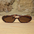 Oval sunglasses with a brown and black tortoiseshell patterned frame and brown tinted lenses, resting on a light wooden surface with a rustic stone wall background.