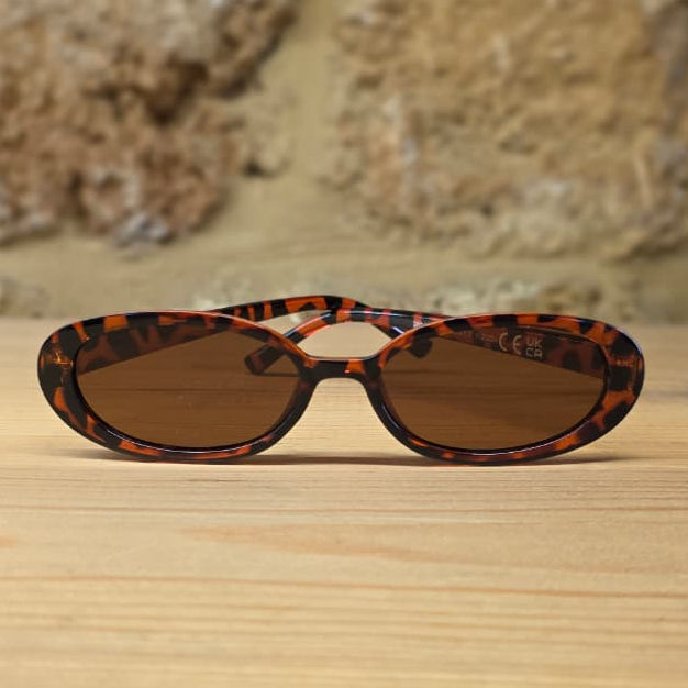 Oval sunglasses with a brown and black tortoiseshell patterned frame and brown tinted lenses, resting on a light wooden surface with a rustic stone wall background.